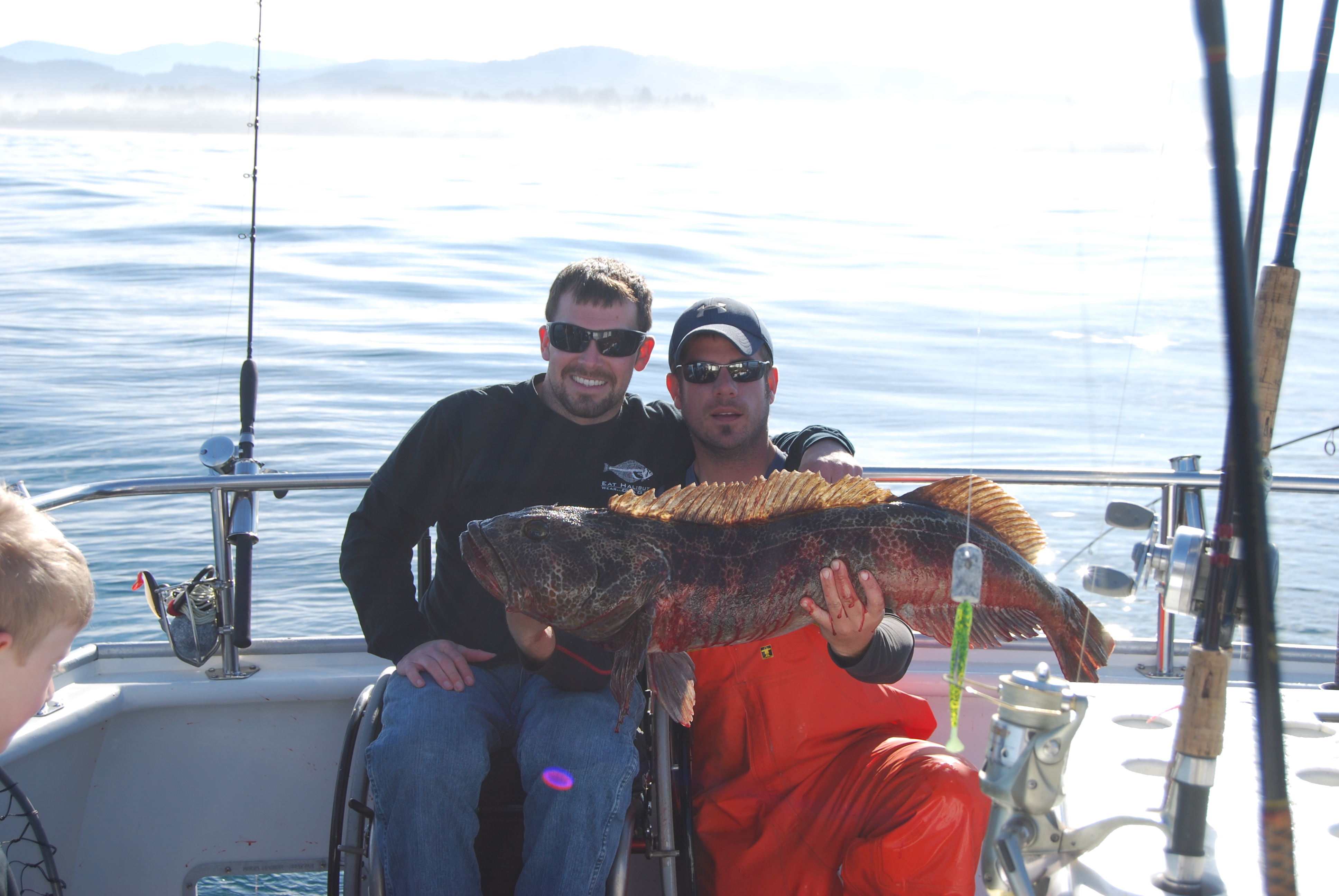 two people holding a lingcod fish aboard a fishing boat with fishing poles, one person is on a wheelchair