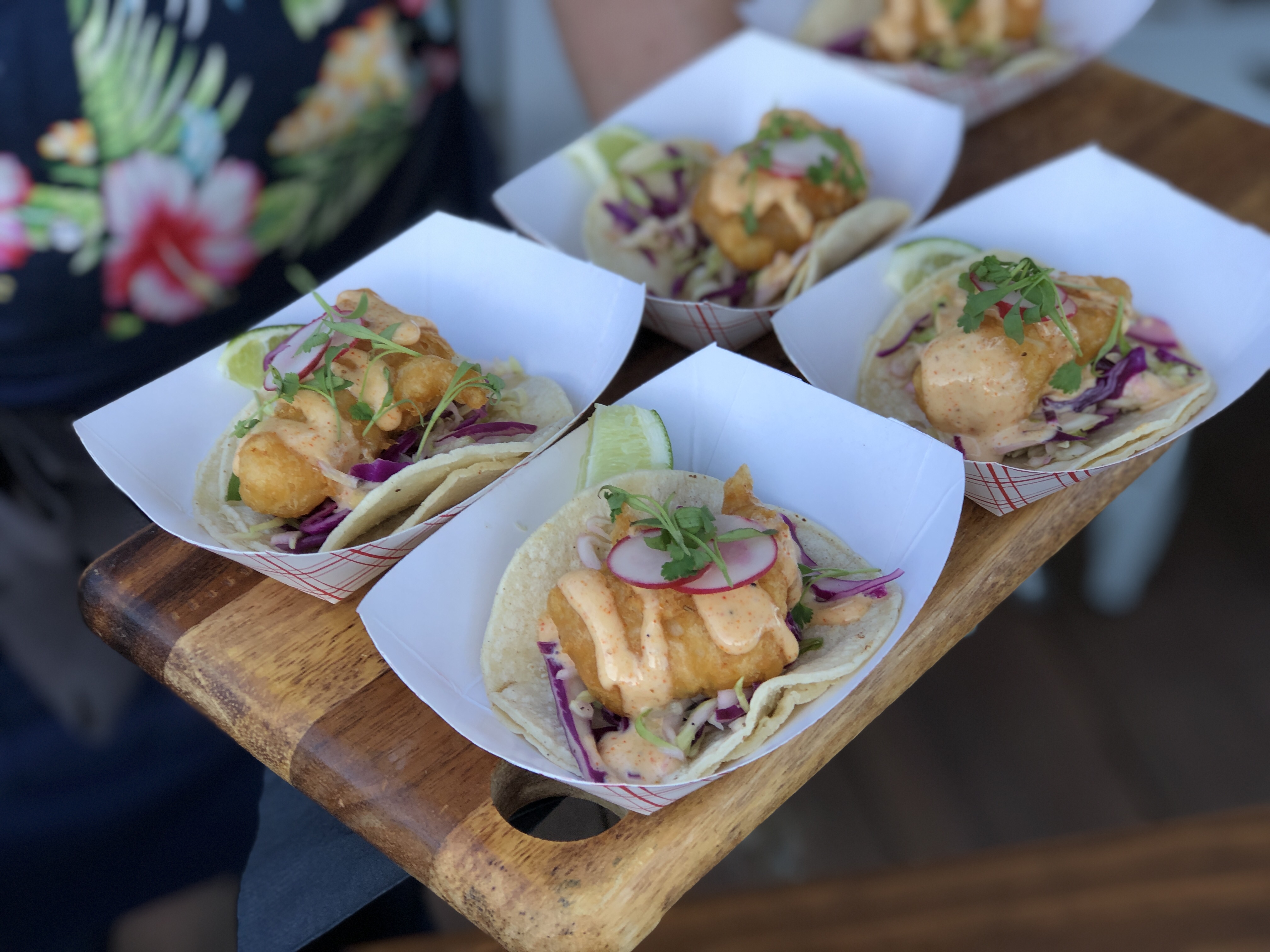 five paper plates with fried fish tacos, each garnished with red cabbage, radish, sauce, cilantro, and lime