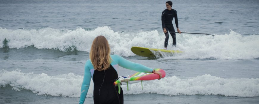 Surfer girl walks into the water carrying a surfboard, while a young man surfs a paddle board on a wave.